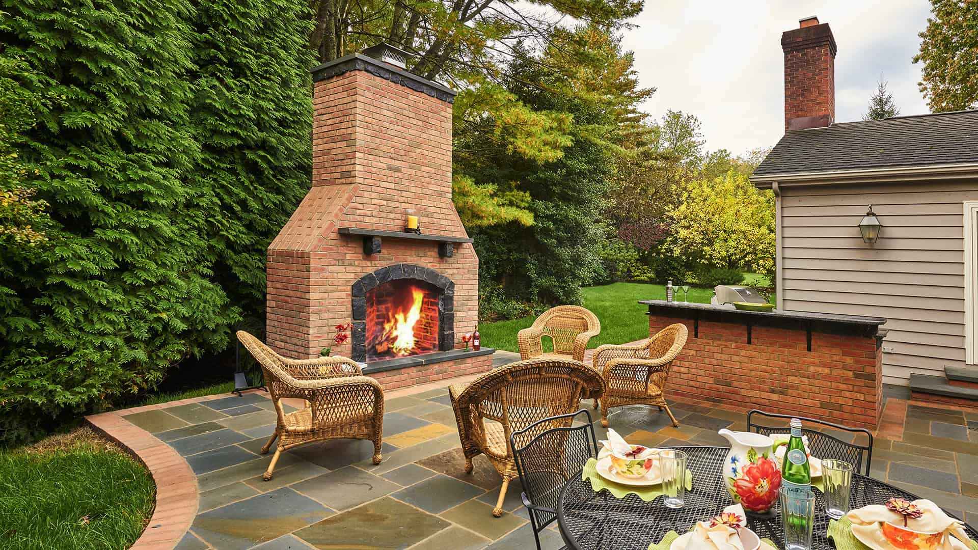 A cozy outdoor patio in Delaware County, PA, features a large brick fireplace with a roaring fire, surrounded by wicker chairs. A black metal table with plates and drinks sits nearby. Lush greenery enhances the inviting atmosphere, showcasing expert hardscaping services.