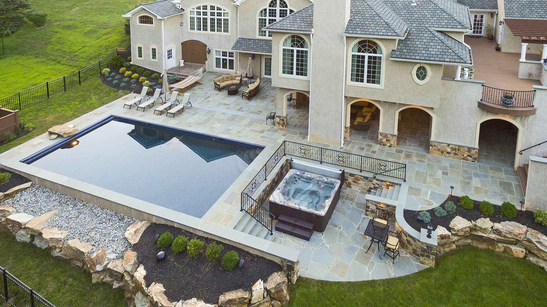Aerial view of a luxurious backyard in Delaware County, PA, showcasing expert landscaping services. The rectangular swimming pool and hot tub are nestled within a stone patio, accompanied by lounge chairs and a seating area. The multi-story house boasts large windows amid lush greenery.