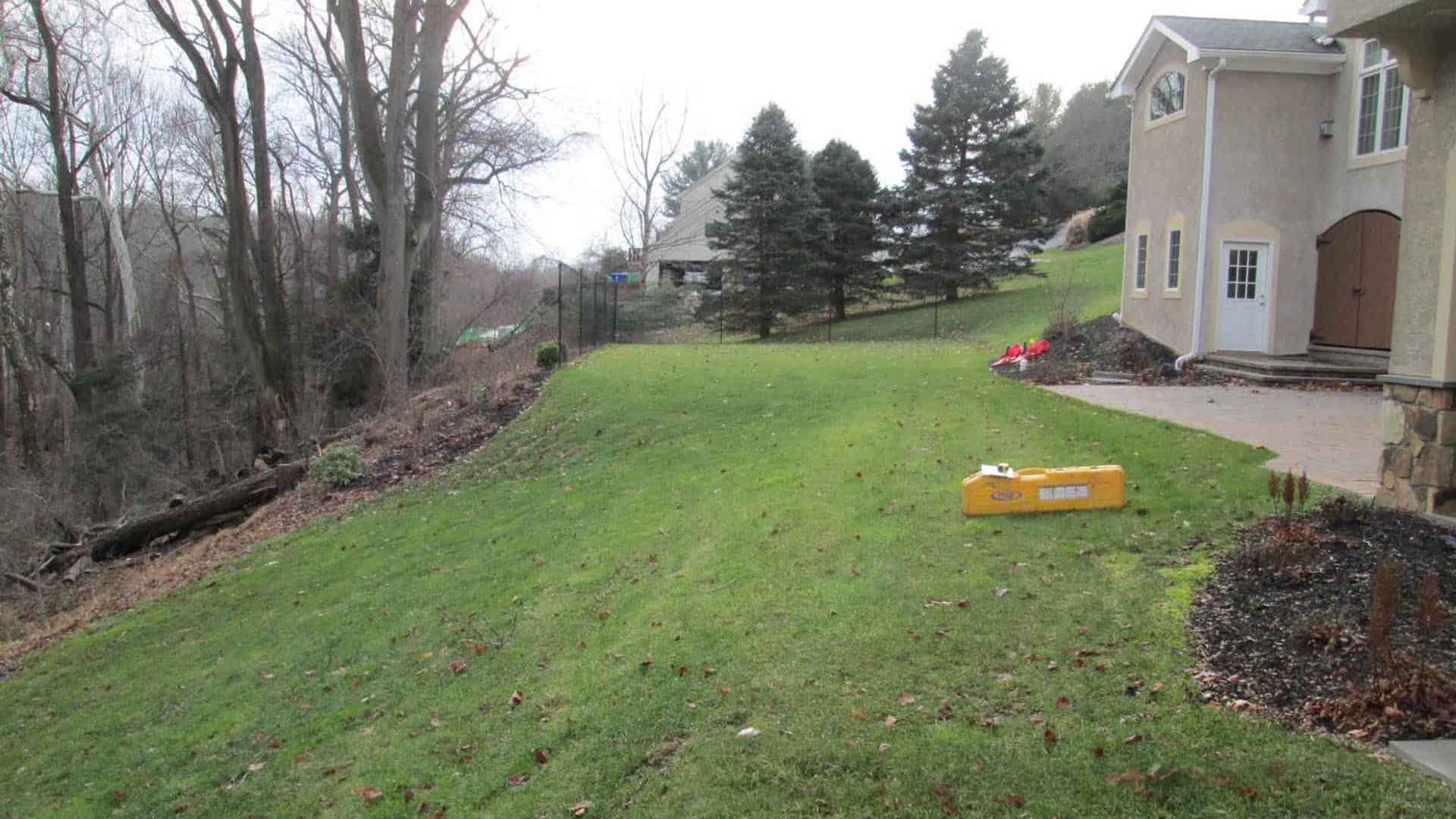 A grassy backyard slopes downward with a few leafless trees on the left and evergreen trees near the center. A beige house stands on the right, and a yellow object lies on the grass. The overcast sky hints at a serene space perfect for outdoor living in Delaware County, PA.