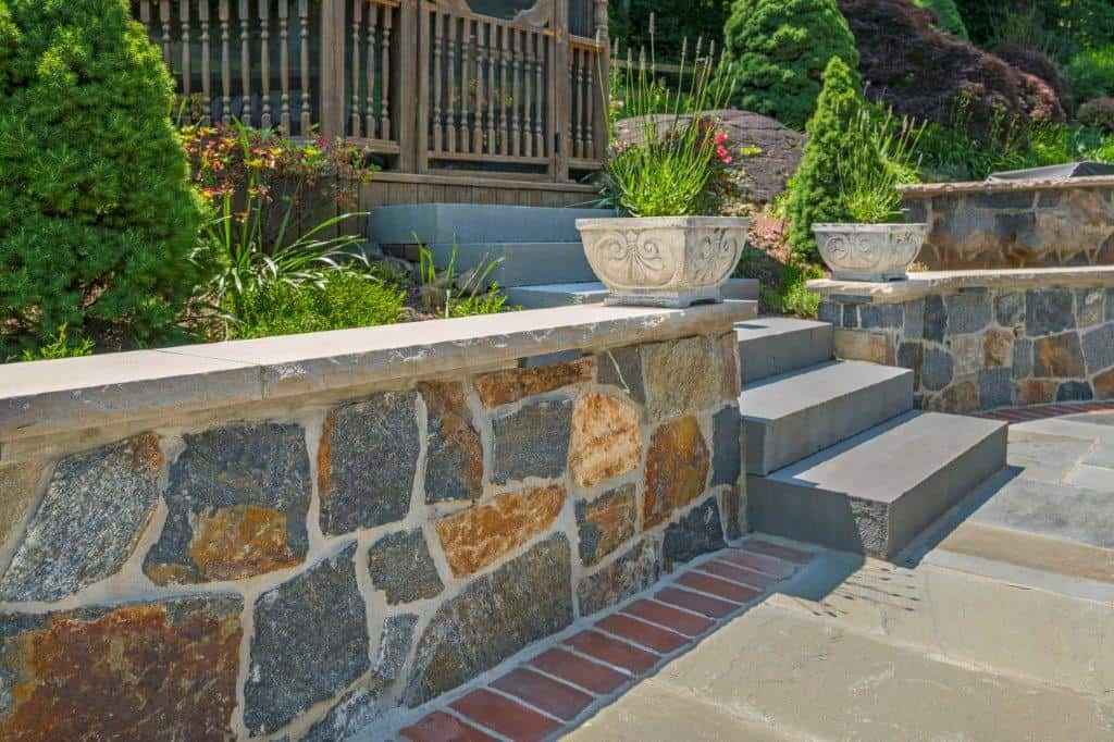 Image of a garden area with a stone retaining wall and stairs leading up to a wooden railing, showcasing expert hardscaping services in Delaware County, PA. Decorative stone planters with green foliage adorn the wall, while lush greenery and paved stones complete this outdoor living space.
