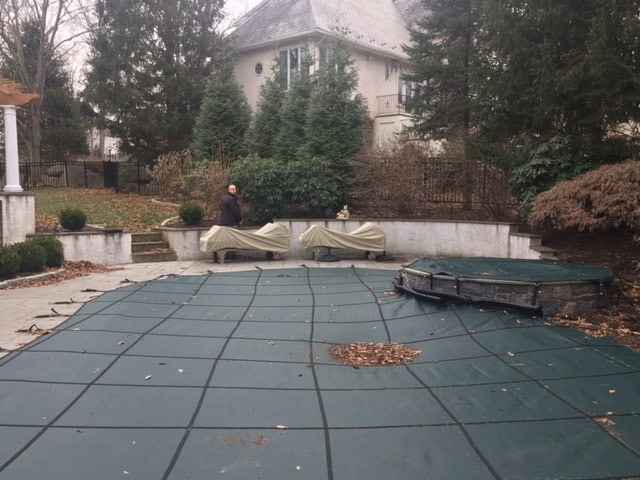 A backyard oasis in Delaware County, PA features a green mesh swimming pool cover surrounded by lush trees and shrubs. Two covered lounge chairs rest near a wall where someone stands. The scene illustrates the art of outdoor living, with a large house providing a picturesque backdrop.