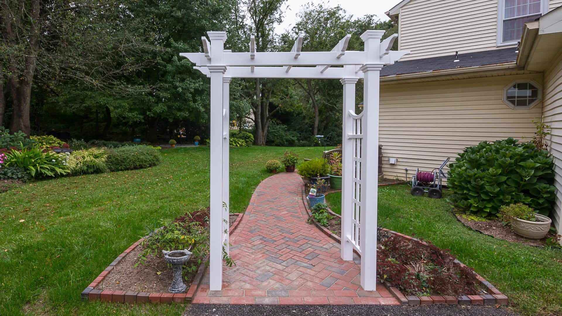 A white garden arbor with a lattice design stands at the entrance of a brick path, exemplifying expert landscaping services in Delaware County, PA. The path is bordered by small plants and leads to a lawn with trees in the background. A beige house is on the right side of the image.