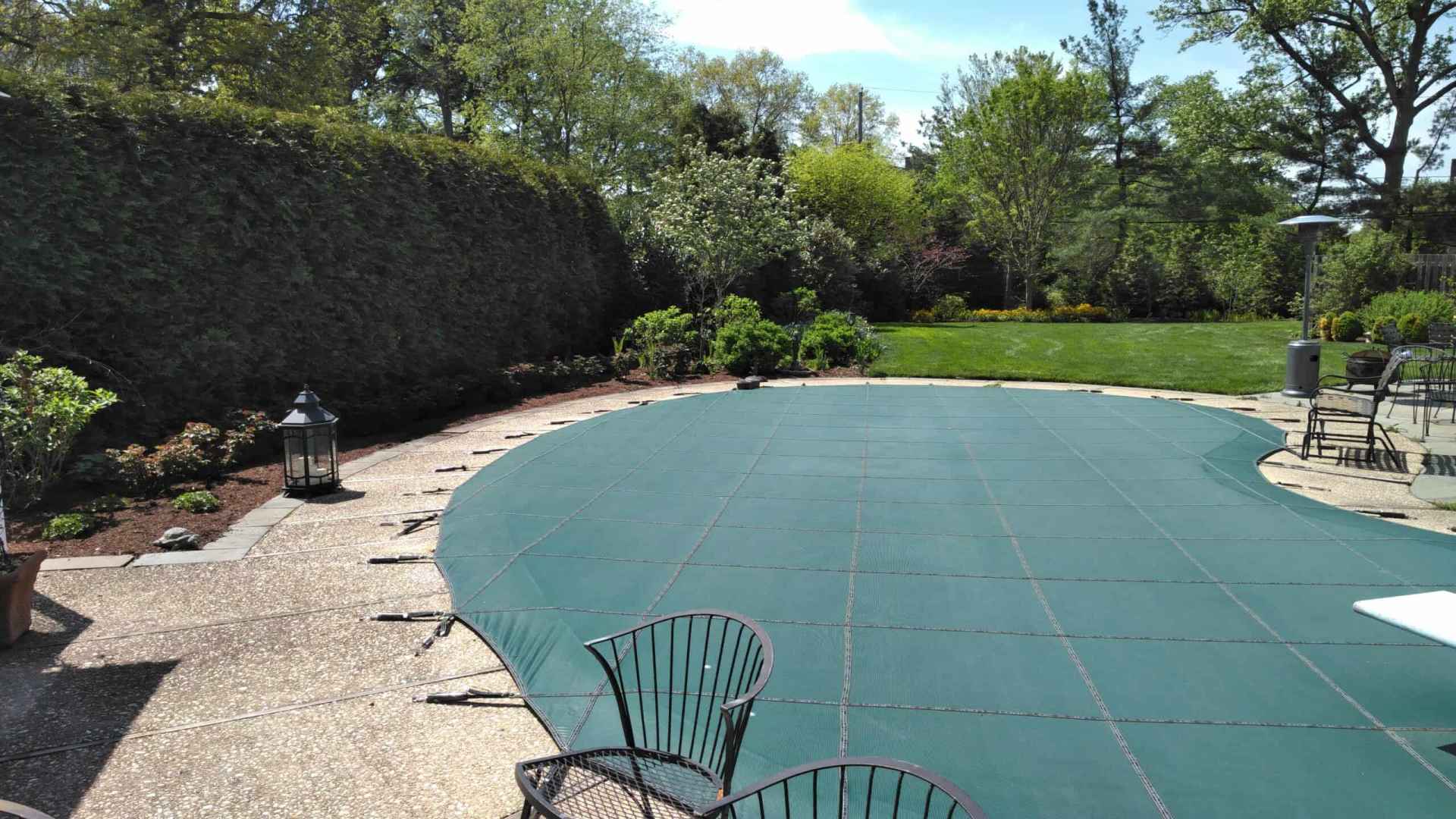 A backyard with a pool covered by a green tarp, surrounded by lush trees and vibrant grass, showcases the charm of outdoor living in Delaware County, PA. Wrought iron chairs and a table grace the patio, while a lantern sits at the edge under the clear sunny sky.