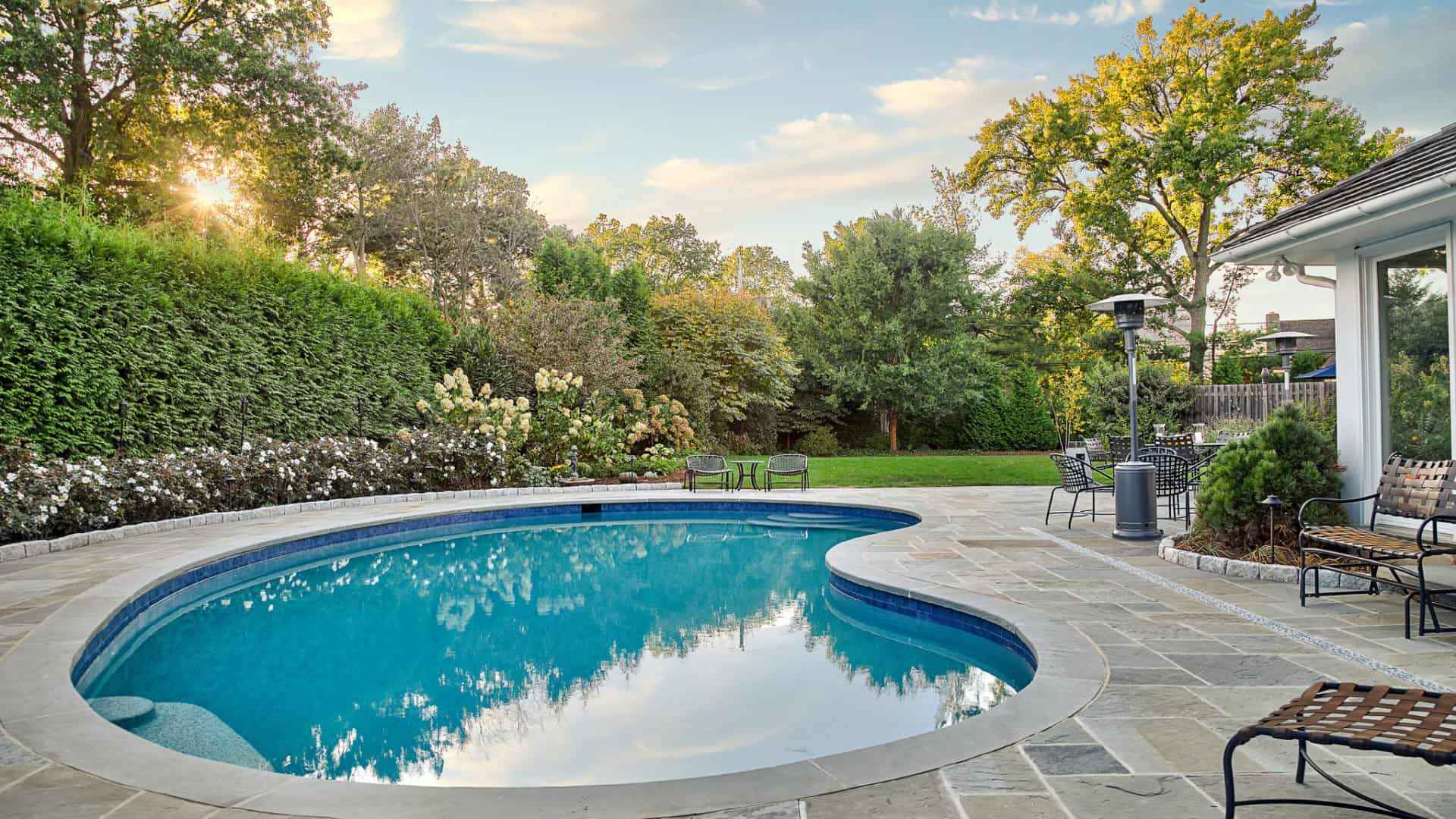A serene backyard in Delaware County, PA features a turquoise kidney-shaped pool surrounded by stone tiles. Green trees and bushes line the background. Patio furniture and a heater are set on the right. The sun sets behind the trees, casting a warm glow over this beautifully hardscaped retreat.