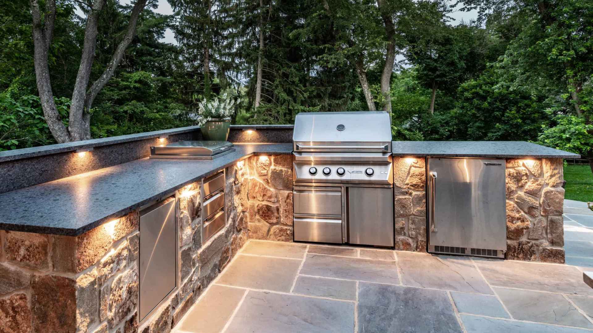 Outdoor living in Delaware County, PA, comes to life with a beautifully designed outdoor kitchen. Featuring stone countertops and cabinets with a stainless steel grill, mini fridge, and storage units on a stone patio. Built-in lighting enhances the space, surrounded by lush greenery.