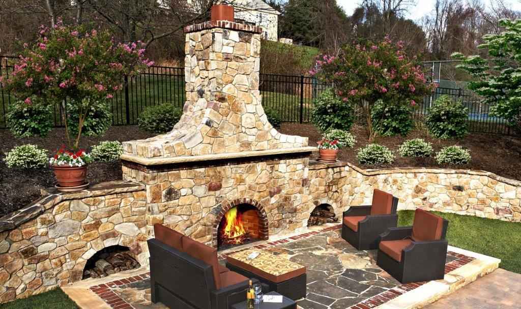 Outdoor patio featuring a stone fireplace and lit fire, perfect for outdoor living in Delaware County, PA. Surrounded by lush trees and plants, four cushioned wicker chairs encircle a rectangular stone table—expertly crafted with our hardscaping services in Delaware County, PA.