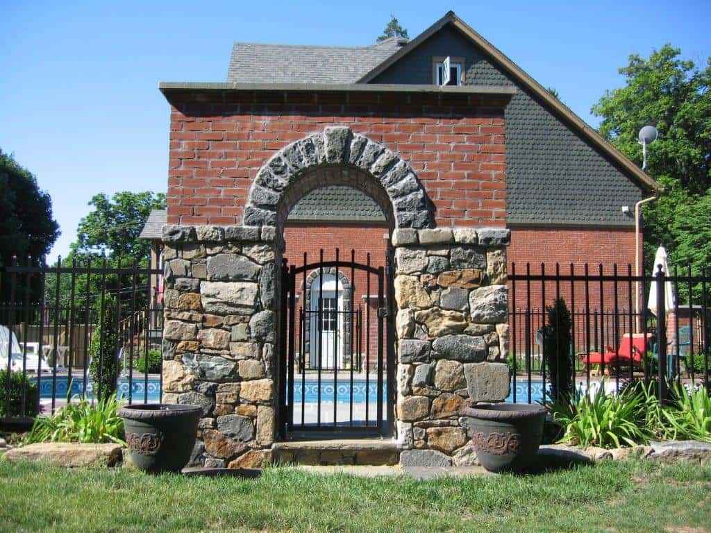 Brick and stone archway with a black metal gate in an elegantly landscaped garden. Beyond, a pool and red brick house with a gabled roof are visible. Two decorative pots flank the archway, embraced by lush greenery—a testament to premier landscaping services in Delaware County, PA.