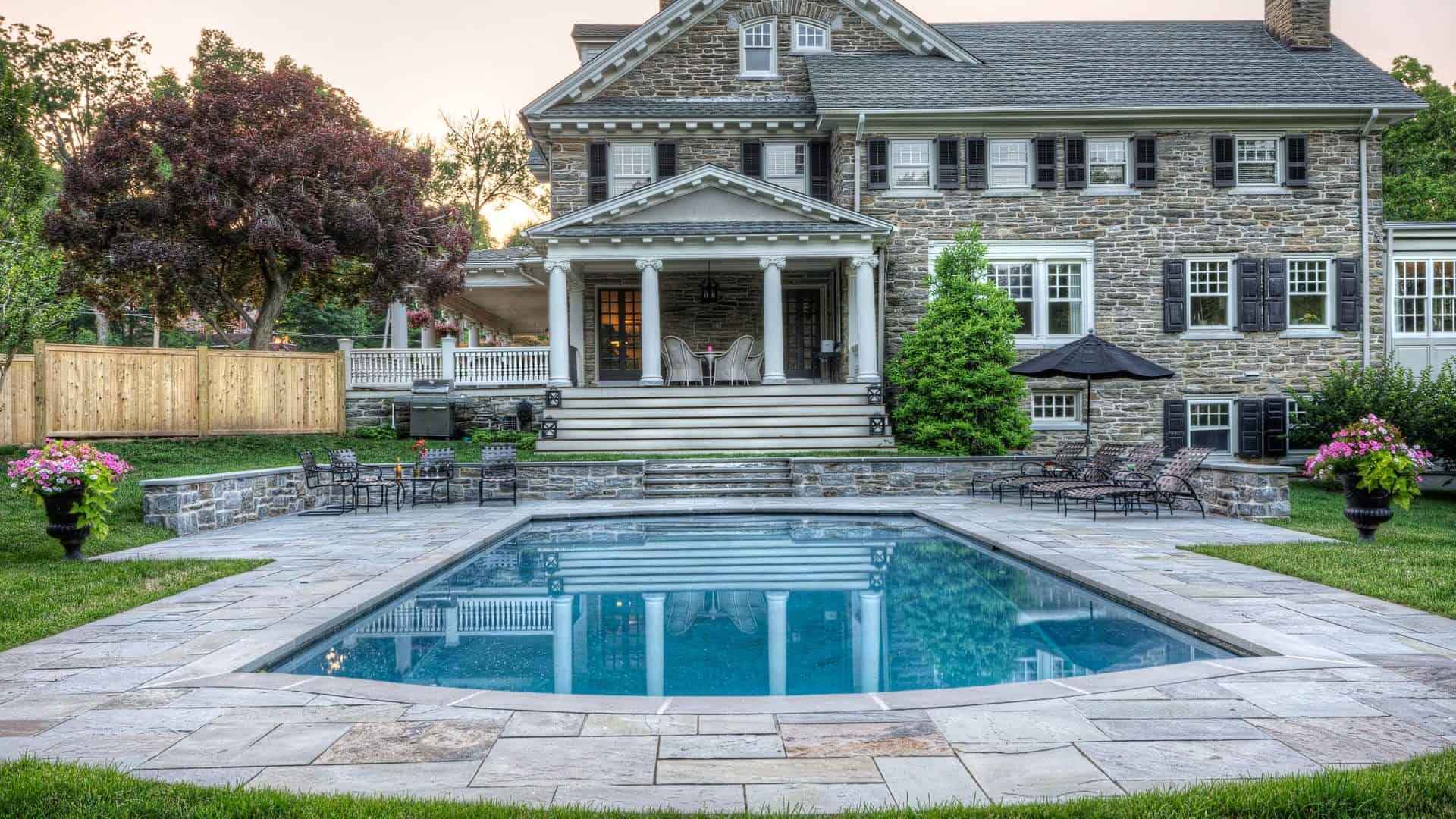 A large stone house with a gabled roof and two-story columns stands behind a rectangular pool. The pool area showcases expert landscaping services from Delaware County, PA, with a stone patio, lush shrubs, and flowers in planters. A wooden fence borders the left side, framed by trees in the background.