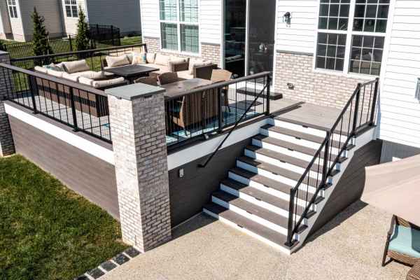 A brick and wood deck with a black railing features outdoor sofas and a wicker dining set, crafted to enhance your outdoor living in Delaware County, PA. Stairs lead down to a concrete patio area surrounded by a grassy lawn, perfectly complementing the house's white exterior with large windows.