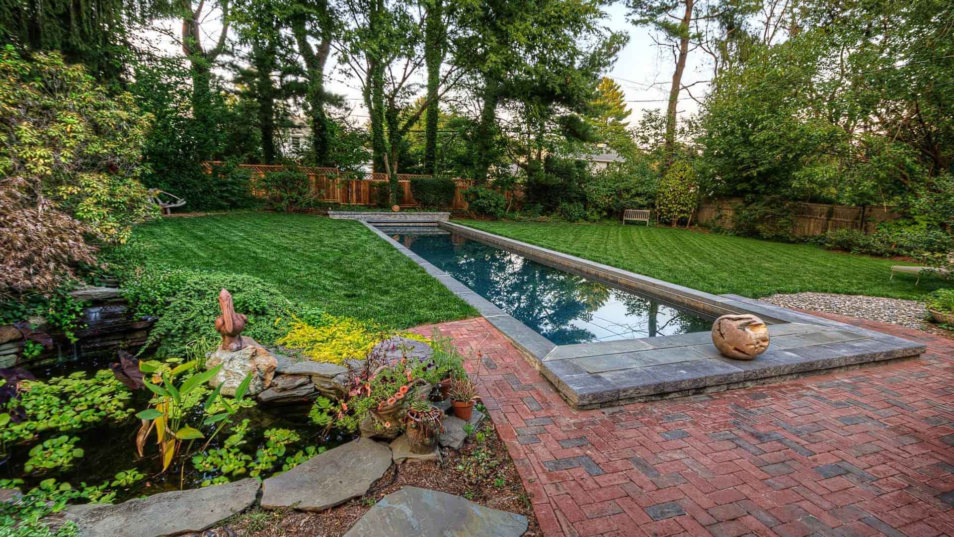 A backyard garden in Delaware County, PA, features a long rectangular pool with stone edging, surrounded by lush green grass and trees. A small stone path with potted plants and a sculpture leads to the pool, complemented by a brick patio area. Consider expert hardscaping services for this outdoor living paradise.