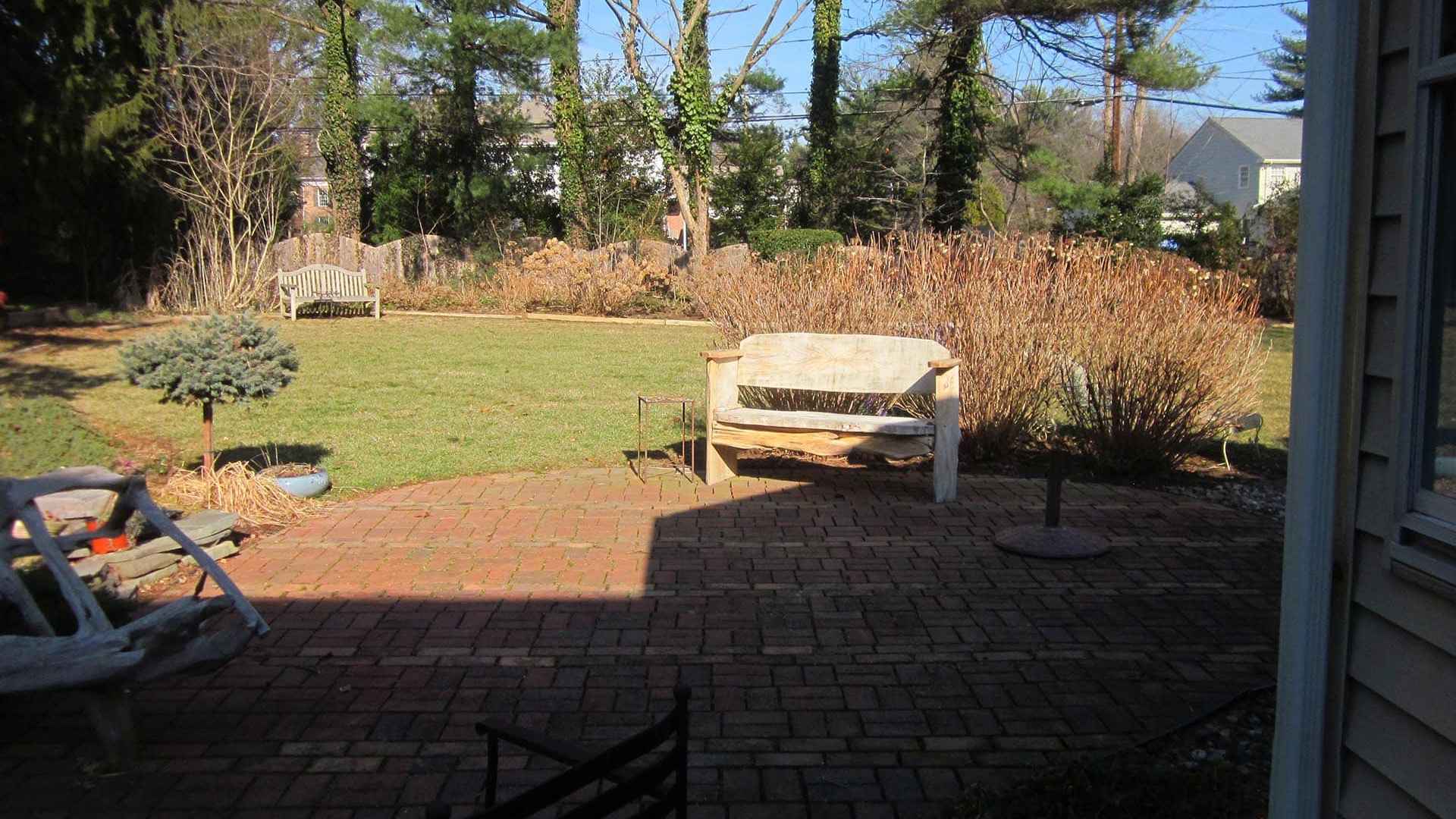 A sunlit backyard with a brick patio showcases outdoor living at its finest in Delaware County, PA. It features garden furniture, including a bench and chairs. Surrounding the patio are lush lawns and trees, with a clear blue sky and a neighboring house visible in the background.
