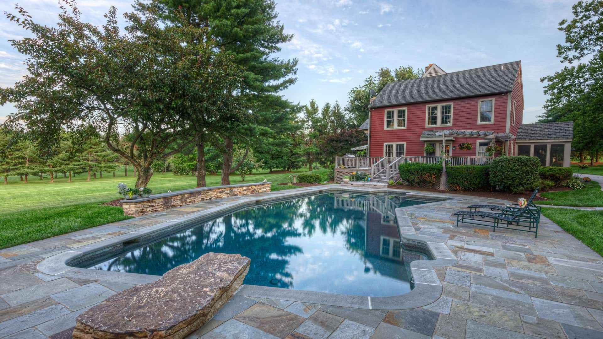 A backyard scene featuring a stone-tiled swimming pool, expertly crafted by renowned hardscaping services in Delaware County, PA. Surrounded by lush green grass and trees, a red two-story house with a porch stands in the background. The sky is partly cloudy, and outdoor chairs invite relaxation near the pool.