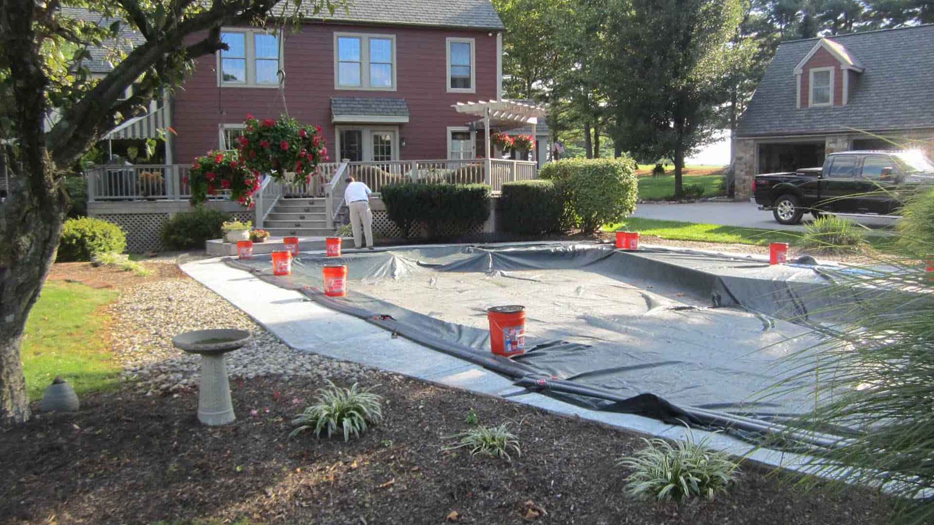 A backyard scene in Delaware County, PA, with a partially covered swimming pool secured by red buckets. A person is working near the pool, surrounded by trees, a stone pathway, and grassy areas. The outdoor living space features a two-story red house and a smaller structure in the background.