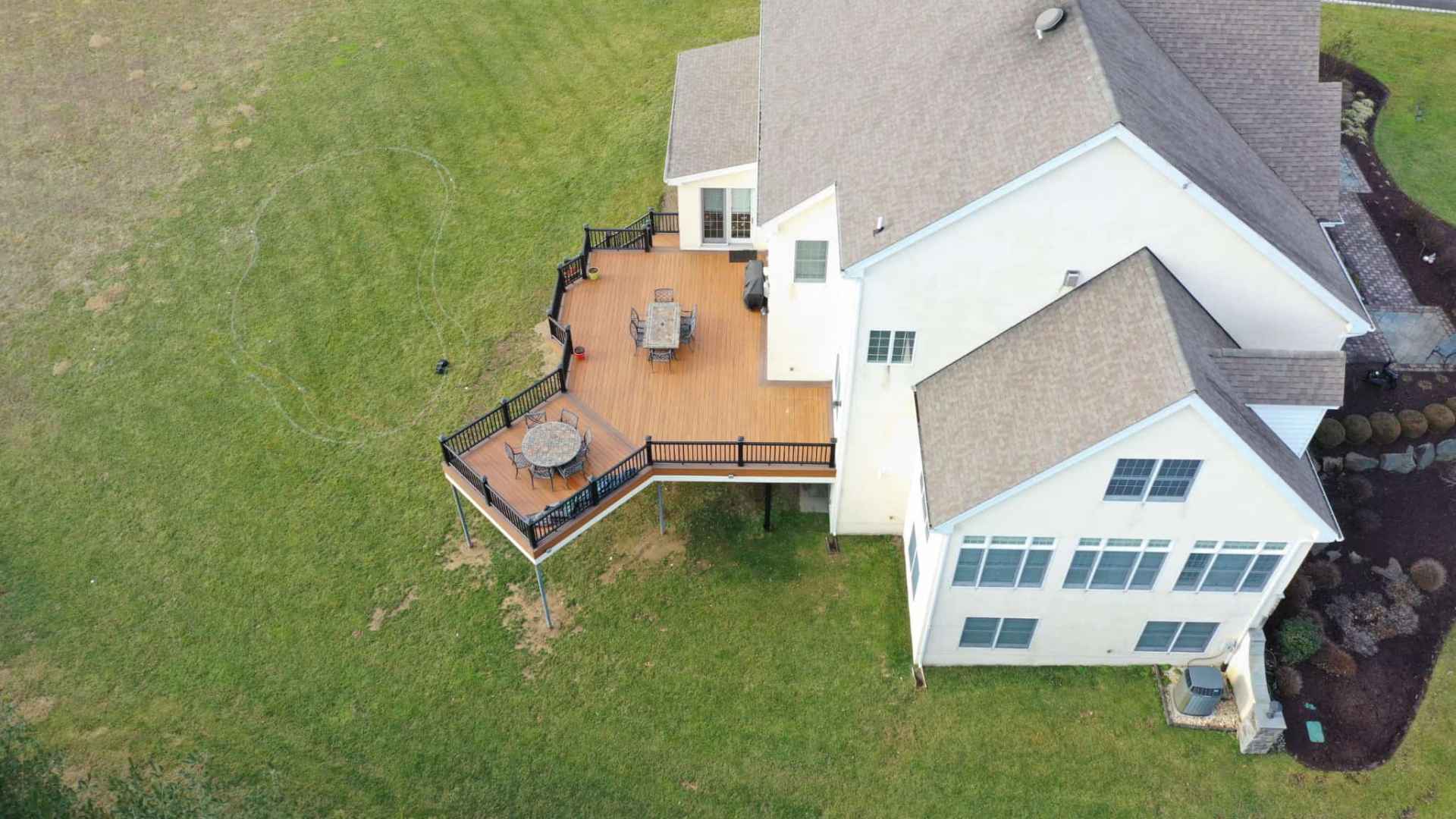 Aerial view of a large house with a spacious wooden deck, perfect for outdoor living in Delaware County, PA. The deck features sleek furniture and overlooks a vast green lawn. Light-colored with numerous windows and a gray roof, the home is complemented by landscaped shrubs and a nearby driveway.