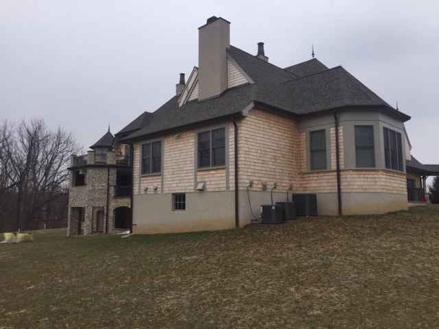 A large, two-story house with beige and gray siding, featuring several chimneys and multiple windows, stands gracefully on a slight hill. With a turret on one side and a grassy yard designed by top landscaping services in Delaware County PA, the scene is serene beneath an overcast sky.