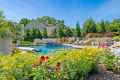 A backyard pool surrounded by vibrant flowers and greenery under a clear blue sky showcases the artistry of outdoor living Delaware County PA. Lounge chairs are positioned on one side, while a house with a beige exterior stands framed by tall trees in the background.