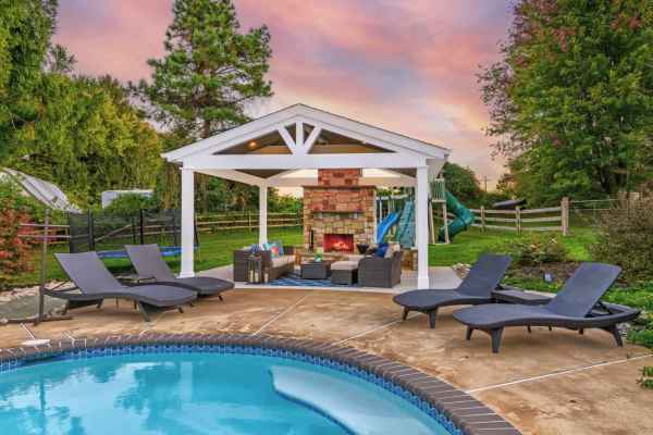 A backyard features a pool with four sun loungers on a patio, perfect for enjoying outdoor living in Delaware County, PA. Nearby, a pavilion with a stone fireplace and seating area is visible. Trees and a slide are in the background, set against a colorful sunset sky.