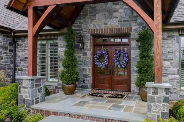 A modern stone house entrance with a wooden gabled roof, exposed beams, and large stone slabs exemplifies the artistry of hardscaping services in Delaware County, PA. The double doors boast decorative wreaths, flanked by tall potted shrubs along a walkway lined with lush greenery.