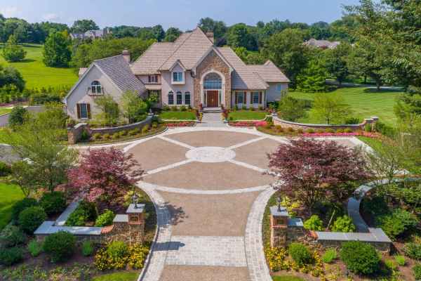 Aerial view of a large, elegant house with a circular driveway and landscaped gardens. The home features a beige exterior with multiple gables, a stone chimney, and a grand entrance. Enhance your outdoor living in Delaware County PA with professional landscaping services for lush greenery and vibrant flowerbeds.