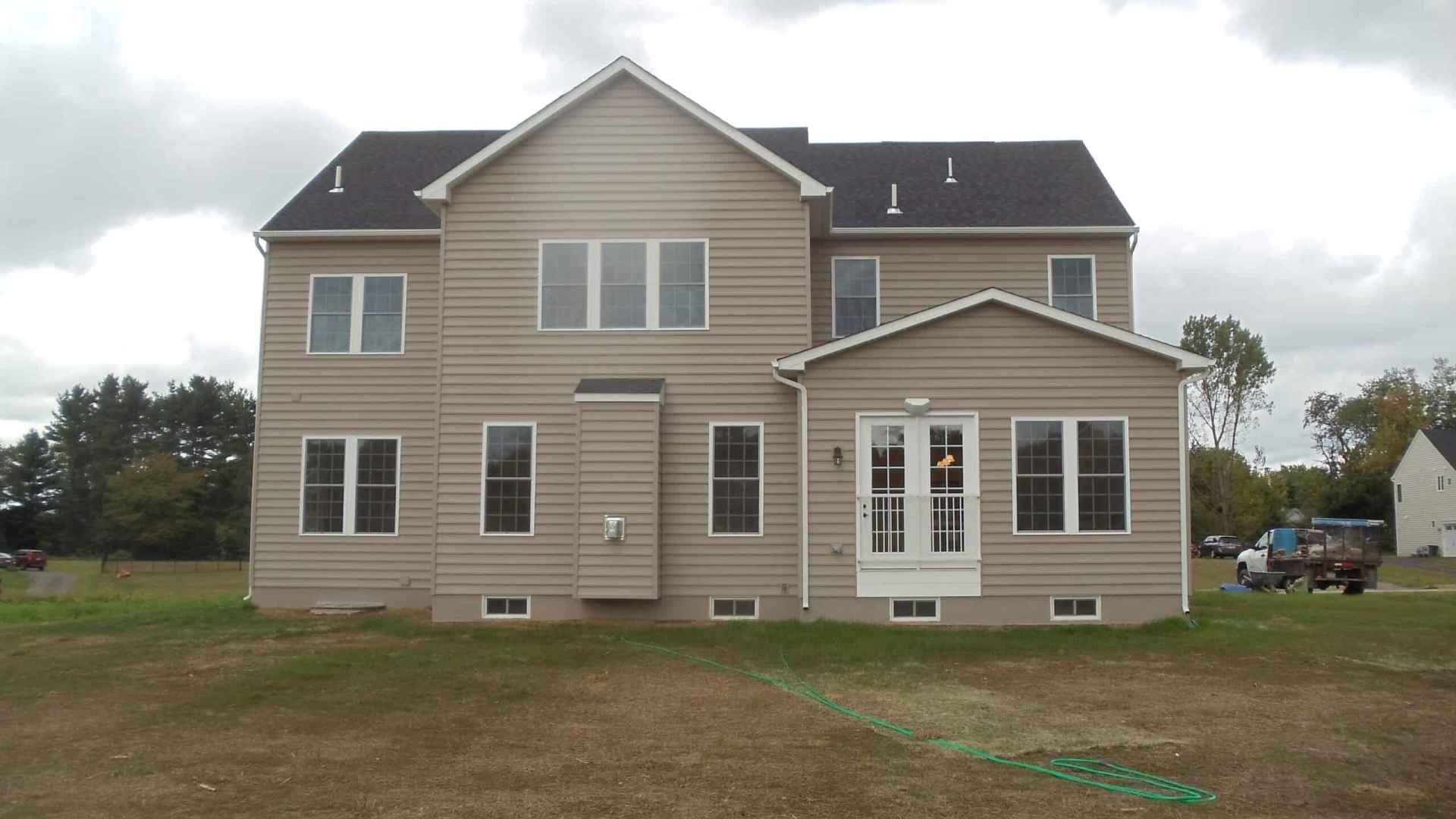 Amidst a cloudy sky, this beige two-story house with multiple windows and a black roof offers an inviting canvas for potential landscaping services in Delaware County, PA. A mostly bare yard with patches of grass awaits transformation beside the green garden hose, while trees frame the scene.