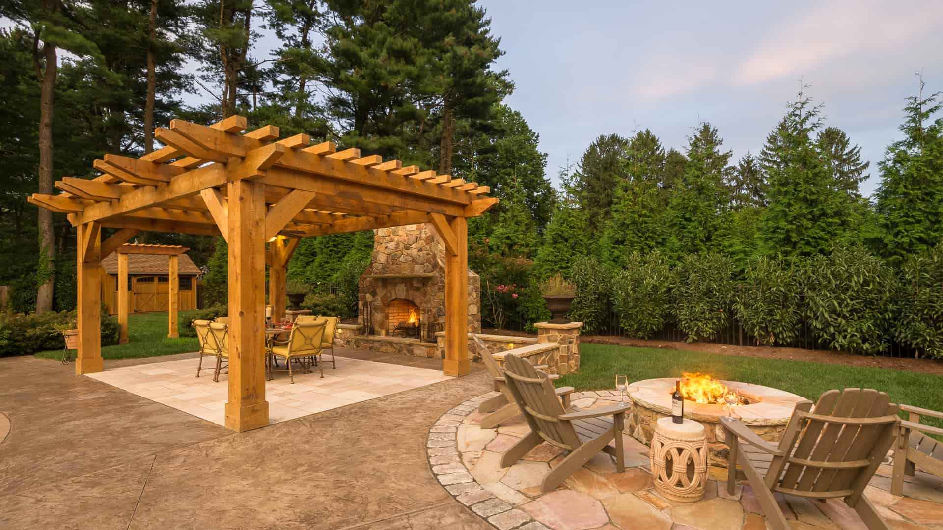A backyard featuring a wooden pergola over a patio with a dining set. Nearby, a stone fireplace and circular fire pit surrounded by Adirondack chairs create the perfect outdoor living space, crafted with expert hardscaping services in Delaware County, PA, all set against tall trees and hedges.