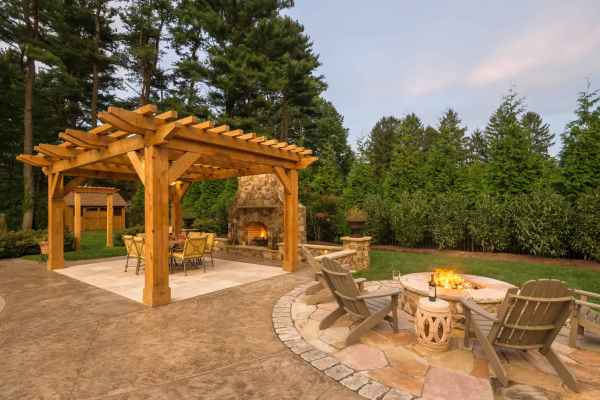 A backyard featuring a wooden pergola over a patio with a dining set. Nearby, a stone fireplace and circular fire pit surrounded by Adirondack chairs create the perfect outdoor living space, crafted with expert hardscaping services in Delaware County, PA, all set against tall trees and hedges.