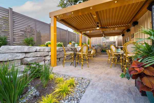 A cozy outdoor patio with yellow chairs and tables nestles under a wooden pergola, offering delightful outdoor living in Delaware County, PA. Bordered by stone walls and lush greenery, slatted fencing ensures privacy while colorful flowers bloom beneath a partly cloudy sky.