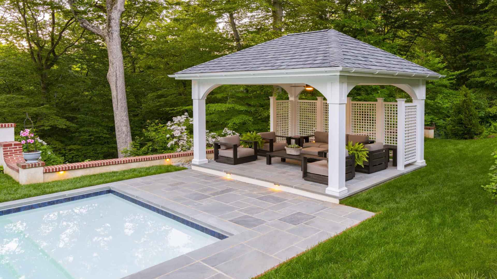 Nestled in a serene setting, this outdoor living space in Delaware County, PA, features a pool with a stone-tiled deck next to a cozy gazebo. Cushioned chairs and a table invite relaxation amid lush greenery and trees, showcasing skilled hardscaping services.