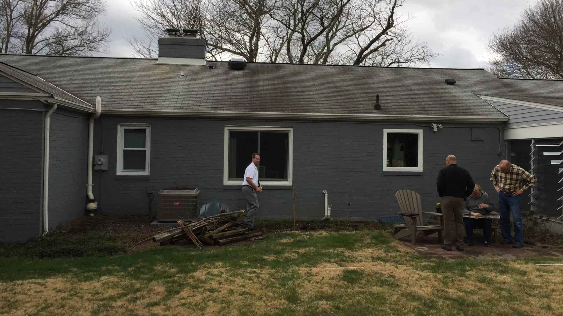 Three people are gathered in the backyard of a gray house with a sloped roof. One stands near stacked wood, while two others examine something on a table. Bare trees and cloudy skies loom overhead, setting the scene for potential outdoor living transformations in Delaware County, PA.