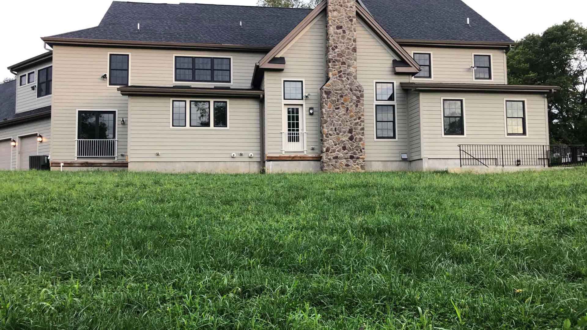 A two-story house with beige siding and a tall stone chimney graces a grassy lawn, showcasing outdoor living at its finest in Delaware County, PA. Numerous windows, a sloping roof, and a small side porch with railing complete the scene, framed by trees in the background.