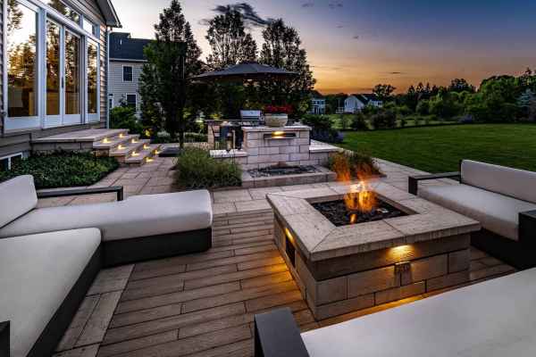 A modern outdoor patio at sunset, showcasing exquisite outdoor living in Delaware County, PA, features white cushioned seating around a lit square fire pit. Illuminated steps lead to a house, with an umbrella shading the grill area. Trees and a lawn frame the scene under a colorful sky.