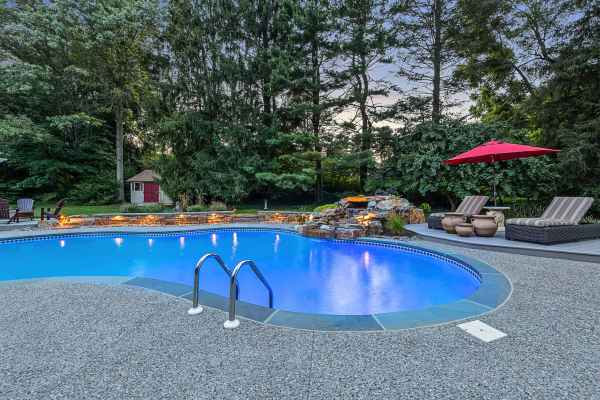 A backyard swimming pool with clear blue water is surrounded by a stone patio, showcasing impeccable outdoor living Delaware County PA appreciates. Two lounge chairs with a red umbrella sit on the right. Trees and a small red shed are in the background, with a waterfall feature cascading into the pool.