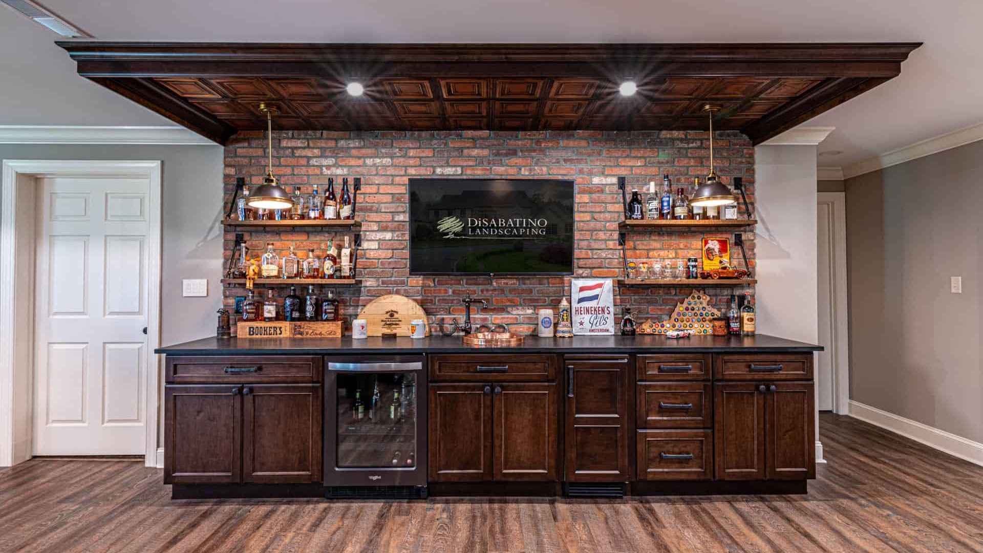 A home bar with a brick accent wall features wooden cabinets and a mini-fridge, seamlessly integrating into the charm of outdoor living. Shelves display liquor bottles and decor. A TV is mounted centrally above the bar, topped with bottles, a sign, and various items.