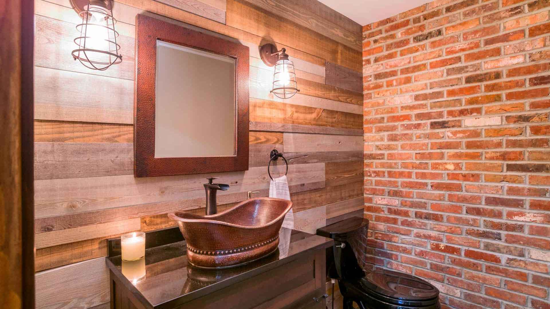 Rustic bathroom with a wooden vanity and copper vessel sink. A rectangular mirror hangs above, flanked by industrial-style fixtures. The walls feature wood paneling, exposed brick, and subtle touches reminiscent of bespoke outdoor living in Delaware County, PA. A lit candle sits on the counter.