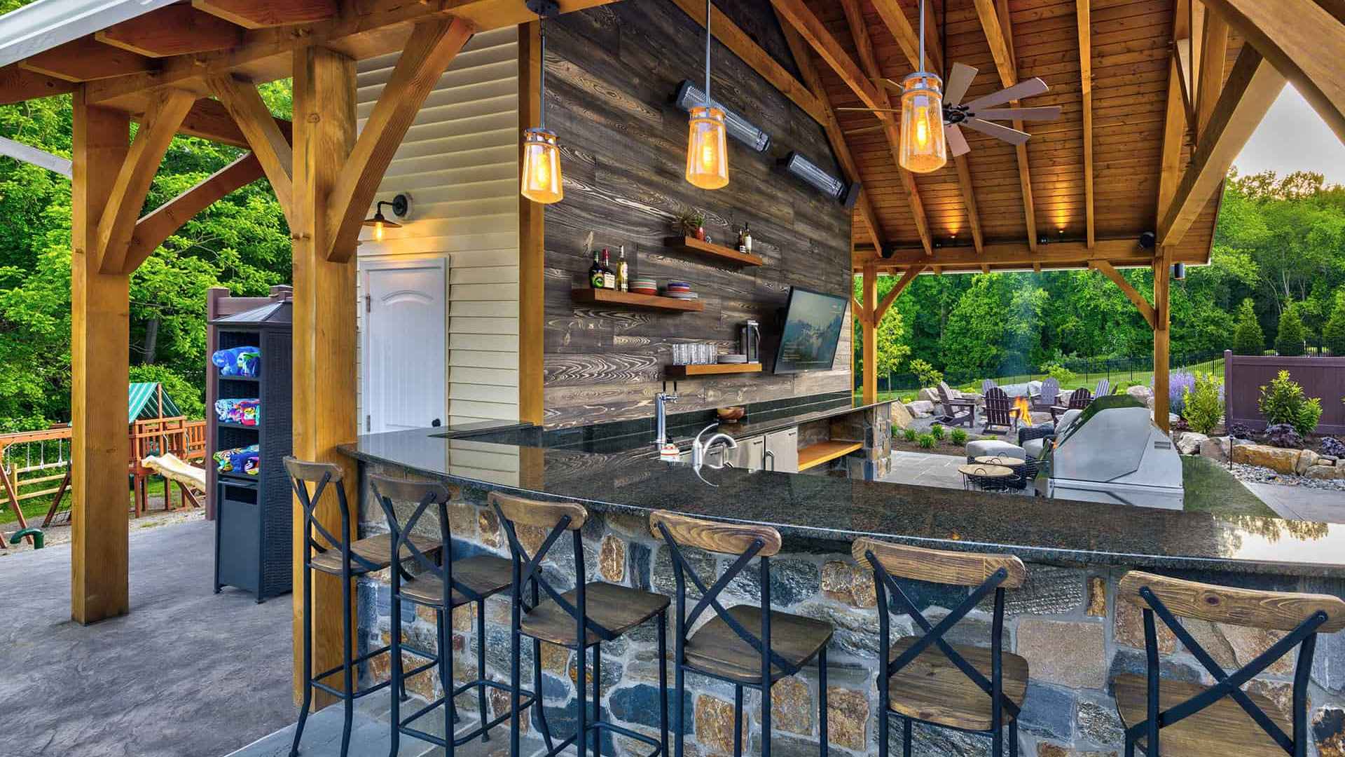 An outdoor bar with a wooden roof and stone counter, surrounded by metal high chairs, embodies the essence of outdoor living in Delaware County PA. Shelves stocked with bottles, a TV, and pendant lights add charm to the space, set against a lush backdrop of trees and an inviting seating area.