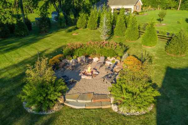 Aerial view of a circular stone patio with a fire pit, crafted expertly by hardscaping services in Delaware County PA. Surrounded by chairs, lush greenery, and trees encircle the patio. A house is visible in the background set within a large, grassy area.