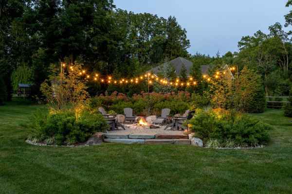 A circular outdoor fire pit area with Adirondack chairs amidst lush greenery and trees showcases exquisite outdoor living in Delaware County, PA. String lights hang above, enhancing the cozy atmosphere. In the background, a house peeks through the trees.