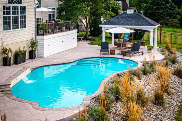 A backyard oasis offers a kidney-shaped swimming pool with clear blue water, framed by beige stone tiles. Discover outdoor living in Delaware County, PA, under a wooden gazebo with a white canopy. Lounge chairs and elegant decorative grasses complete this serene retreat.