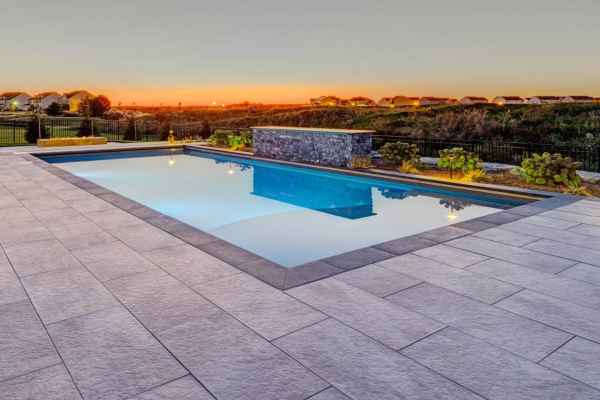 A rectangular swimming pool with clear water reflecting the sunset and a modern water feature at one end. Surrounded by stone tiles from top-notch hardscaping services in Delaware County, PA, it overlooks a grass field and distant houses under a colorful evening sky.