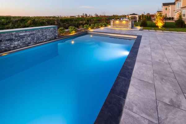 A modern outdoor swimming pool with clear blue water is surrounded by a sleek stone deck, exemplifying top-tier hardscaping services in Delaware County, PA. In the background are a fire pit, a few bushes, and a house under a clear sky, suggesting a serene, early evening setting.