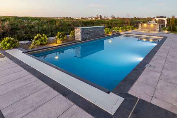 A modern rectangular swimming pool, surrounded by wide gray tiles, reflects the evening sky. In the background, a stone wall with lights enhances the landscaped area featuring greenery and a distant gazebo. Perfect for outdoor living, this Delaware County, PA oasis elevates any hardscape design.