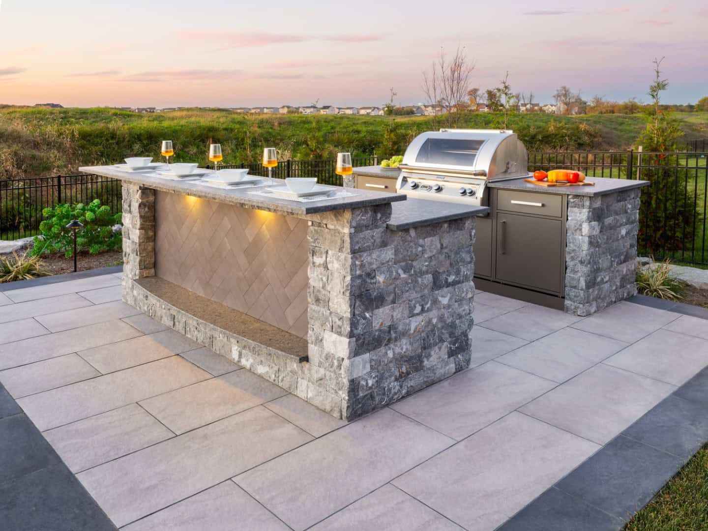 Discover the charm of outdoor living in Delaware County, PA, with a patio kitchen that boasts a stone island complete with plates, glasses, and a built-in grill. Surrounded by lush landscaping services and offering breathtaking sunset views, it's your perfect escape.