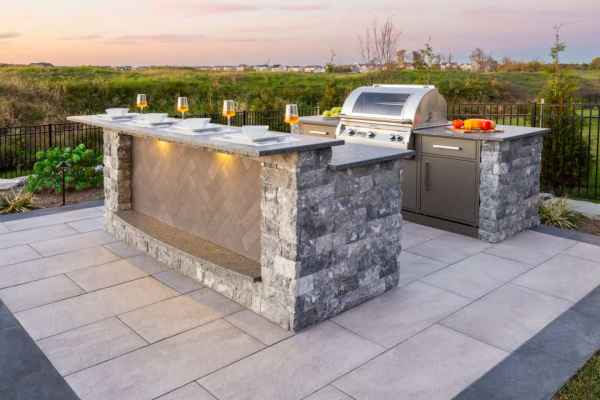 Discover the charm of outdoor living in Delaware County, PA, with a patio kitchen that boasts a stone island complete with plates, glasses, and a built-in grill. Surrounded by lush landscaping services and offering breathtaking sunset views, it's your perfect escape.