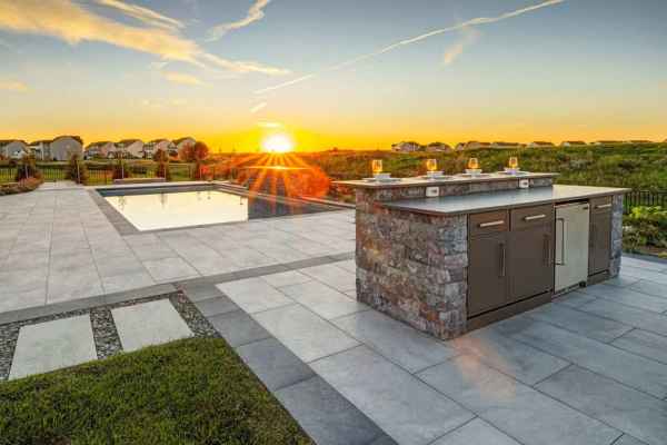 Outdoor patio with a stone kitchen island and stainless steel appliances at sunset. A rectangular pool reflects the sky in this outdoor living haven in Delaware County, PA. The area features light stone tiles bordered by grass, offering hardscaping services with a scenic horizon view.