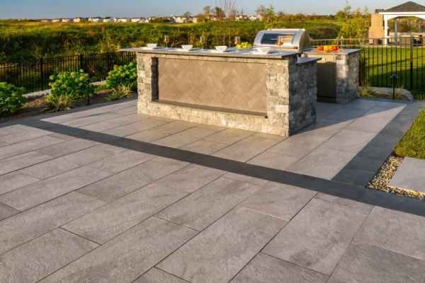 Outdoor living in Delaware County, PA, elevated by a stone island and built-in grill on a spacious patio with large gray tiles. Green bushes and a grassy field surround the area, with houses and a gazebo visible in the background under a clear blue sky.