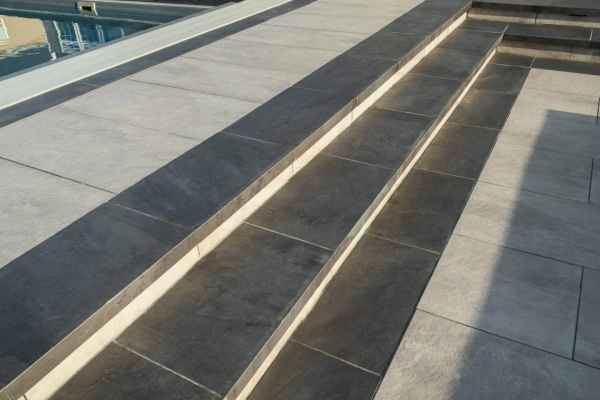 Close-up of a modern outdoor space in Delaware County, PA, featuring light and dark gray stone tiles. The design includes multiple levels with steps leading to an area, possibly a pool, which has a protective cover. Shadows cast across the tiles create a geometric pattern.