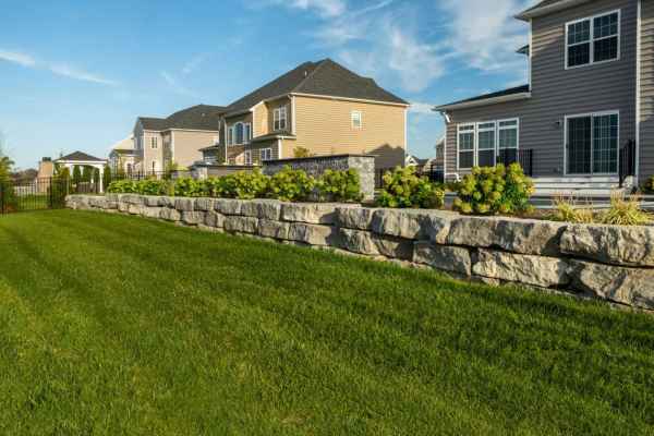 A row of suburban houses with well-maintained lawns is visible. In the foreground, a neatly arranged stone retaining wall supports a garden with green shrubs. The scene showcases expert landscaping services in Delaware County, PA, under a sky that's blue with scattered clouds.