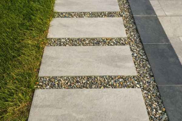 A modern garden pathway with square concrete tiles set in small, gray pebbles. The path is bordered by green grass on one side and a larger paved area on the other, enhanced by decorative plants—ideal for those seeking expert landscaping services in Delaware County, PA.
