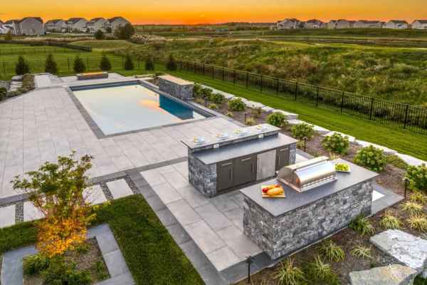 A modern backyard boasts a rectangular pool on a tiled patio. Nearby, an outdoor kitchen with grill and counter space enhances the charm. Expertly crafted by hardscaping services in Delaware County PA, it’s all set under a vibrant orange sunset sky, framed by lush shrubs and grass.