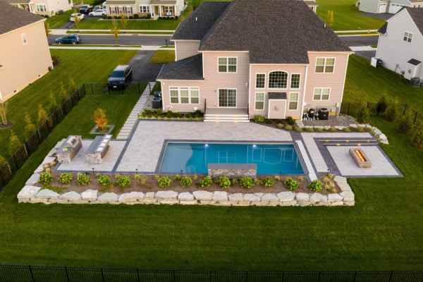 Aerial view of a suburban backyard featuring a modern house with a large pool, showcasing expert landscaping services in Delaware County PA. The yard boasts neatly arranged stone tiles, a fire pit area, landscaped greenery, and outdoor seating. Surrounding houses and road are visible in the background.